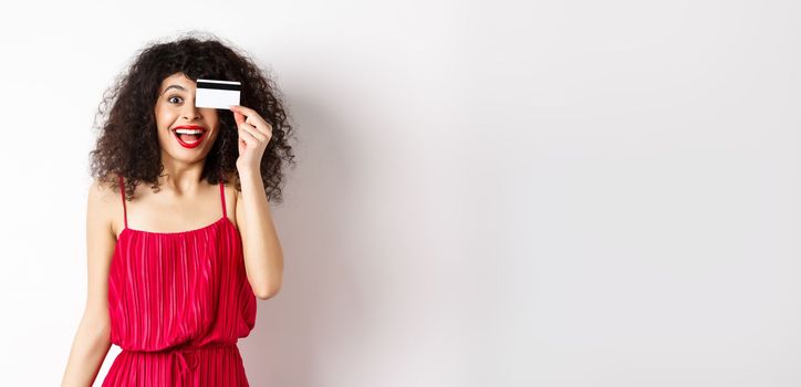 Shopping. Excited curly girl in red dress, showing plastic credit card and scream from joy, amazed with discounts and prices, standing against white background.