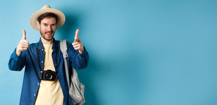 Handsome tourist going on summer vacation with camera and backpack pointing fingers at camera and inviting you, standing on blue background.