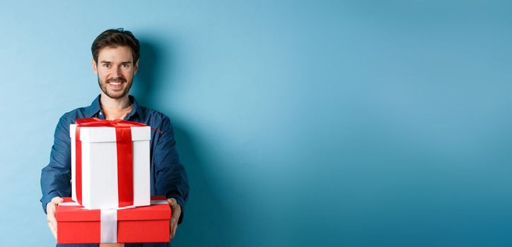 Happy valentines day. Handsome man giving girlfriend presents, holding gift boxes and smiling, standing over blue background.