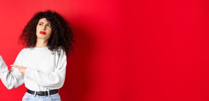 Skeptical frowning girl with curly hair, pointing and looking left hesitant, standing upset on red background.