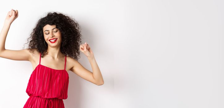 Carefree woman with curly hair, dancing at party, wearing red dress, relaxing with music, standing on white background.