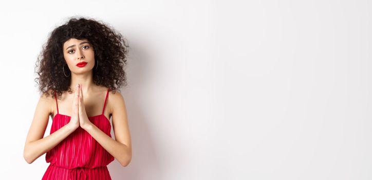 Lady with curly hair, wearing dress and begging for help, holding hands in pleading gesture, asking for favour, standing on white background.