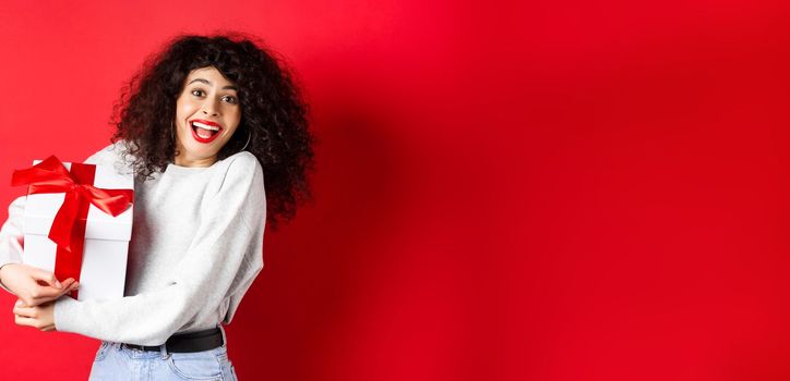 Valentines day. Happy girlfriend holding gift box from secret admirer, looking romantic at lover, receive present, standing on red background.