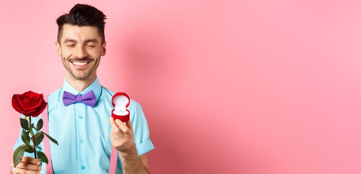 Valentines day. Cute boyfriend making wedding proposal, showing engagement ring in small box and red rose, express love, standing over pink background.