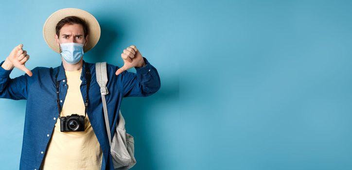 Covid-19 and summer holidays concept. Disappointed tourist in medicla mask frowning, showing thumbs down, negative feedback about travel, standing on blue background.