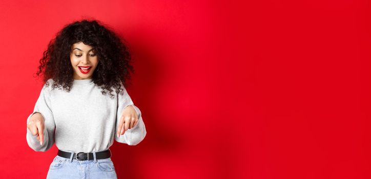 Dreamy beautiful woman with curly hair, pointing and looking down excited, checking out promo, standing against red background.