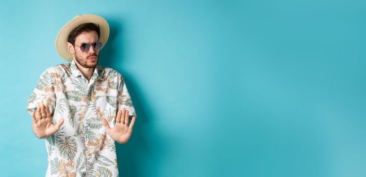 Alarmed tourist asking to stay away, step back from something cringe, showing rejection gesture, standing in straw hat and hawaiian shirt, blue background.