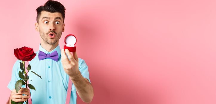 Valentines day. Romantic funny guy going to make wedding proposal, asking to marry him, holding red rose and looking at engagement ring with excitement, pink background.