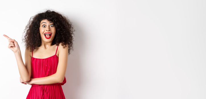 Excited smiling woman with curly hair and makeup, wearing elegant red dress, scream surprised and pointing finger left at logo, standing on white background.