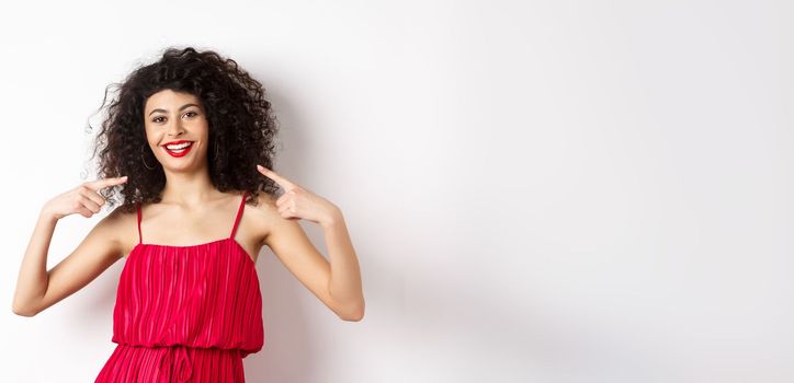 Beautiful woman with curly hairstyle, makeup and red dress, pointing at herself, smiling with white teeth, standing on white background.