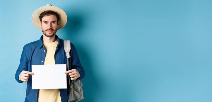 Image of smiling guy in straw hat backpacking, hitchhiking with piece of paper, travelling abroad on summer vacation, blue background.