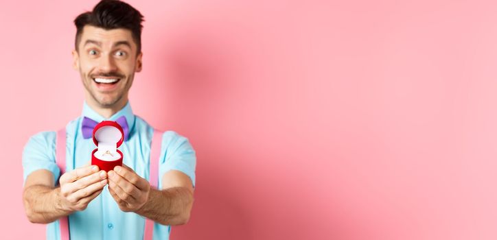 Valentines day. Romantic young man in bow-tie stretch hands with engagement ring and smiling, asking to marry him, making proposal, standing over pink background.