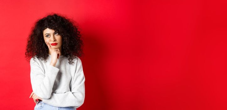 Grumpy young woman with curly hair, looking annoyed or bored at empty space, standing pensive and sad on red background.