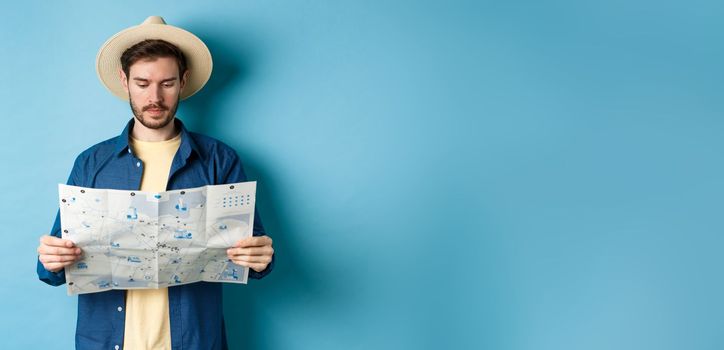 Handsome tourist with summer hat reading map, looking for sightseeing on vacation, standing on blue background.
