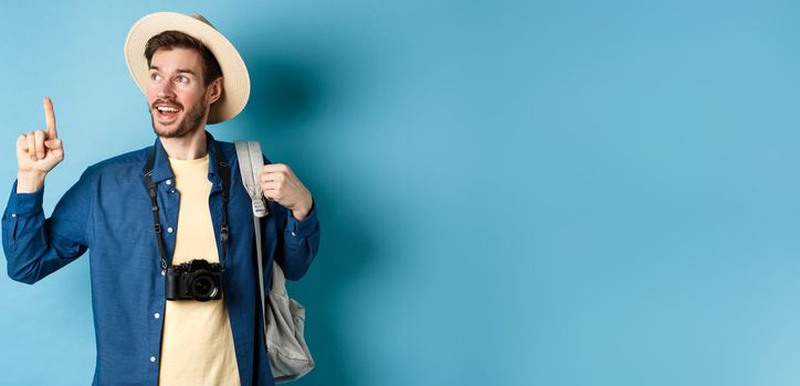 Handsome tourist looking and pointing up, travelling on summer vacation with backpack and camera, standing on blue background.