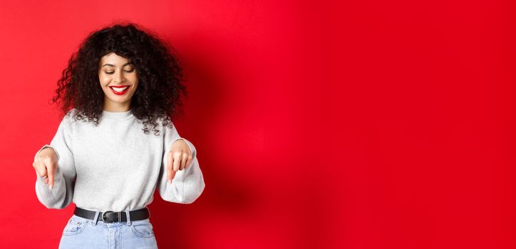 Modern woman with curly hairstyle and red lips, pointing fingers down and smiling happy, showing advertisement on empty space, studio background.