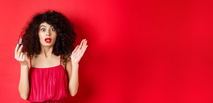 Beauty and makeup concept. Excited woman with curly hair, gasping as looking at camera, holding red lipstick, standing in dress on white background.