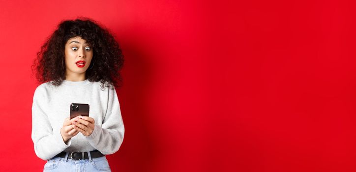 Shocked woman stare at smartphone screen with popped eyes, reading strange message, standing on red background.