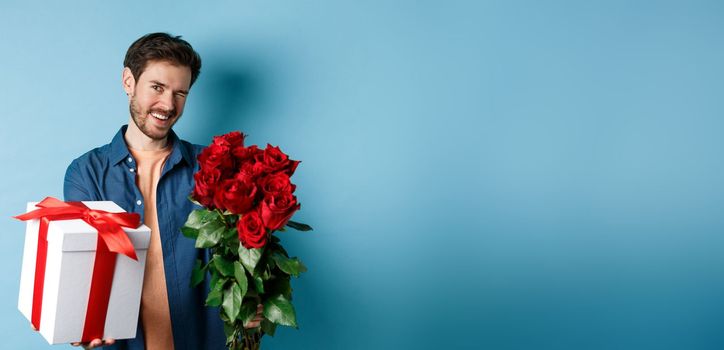 Love and Valentines day concept. Happy guy winking and smiling at camera, presenting a gift and bouquet of flowers to girlfriend, standing over blue background.
