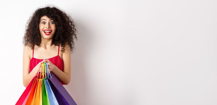 Cheerful young woman in stylish red dress, holding shopping bags and smiling, buying during promo offers, standing over white background.