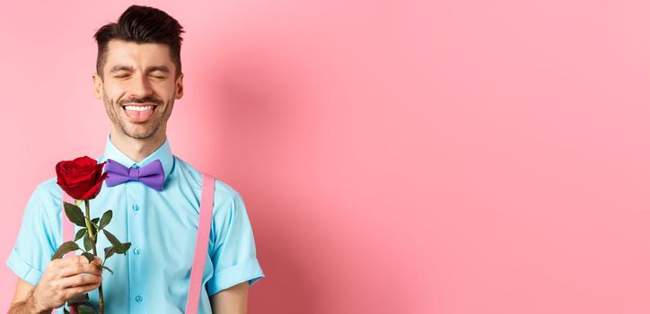 Happy man showing tongue and smiling, holding red rose for girlfriend on Valentines day, enjoying romantic date, pink background.