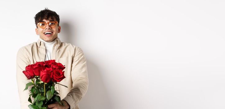 Man in love holding red roses and looking tenderly at camera, staring at lover with happy face, celebrating Valentines day with girlfriend, standing over white background.