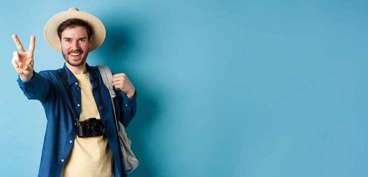 Handsome positive guy tourist showing peace sign on summer vacation, enjoying tropical holidays under sun, wearing straw hat, holding backpack and camera, blue background.