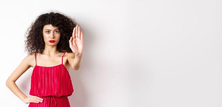 Serious and confident woman in red dress and makeup stretch out hand, tell to stop, prohibit and forbid something, standing over white background.