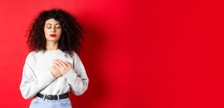 Image of calm young woman with curly hairsty, close eyes and holding hands on heart, keeping warm memories, feeling nostalgic, standing on red background.