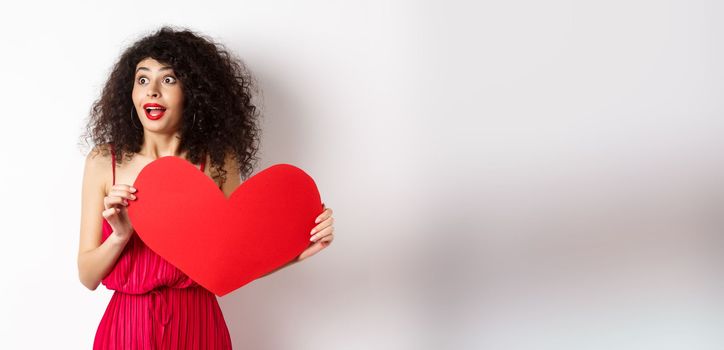 Valentines day. Young woman looking surprised left, seeing something amazing on lovers day, found true love, holding big red heart on chest, white background.