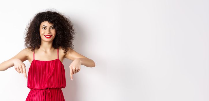 Attractive female model in red dress and makeup, pointing fingers down and smiling, showing advertisement, standing over white background.