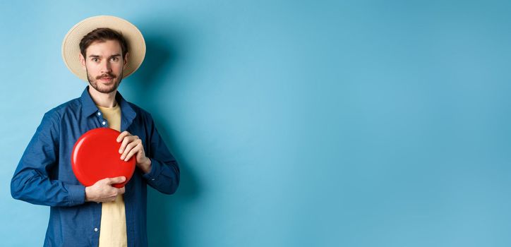 Young man throwing frisbee, playing on summer vacation, standing in straw hat on blue background.