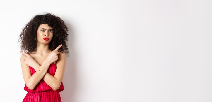 Doubtful and sad young woman in red dress, pointing fingers sideways and sulking, cant decide between products, need help with choice, standing over white background.