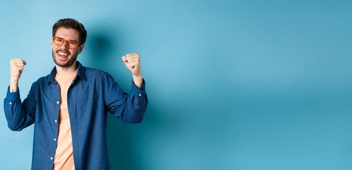 Cheerful young man celebrating, raising hands up and saying yes, wearing eyewear, standing on blue background.