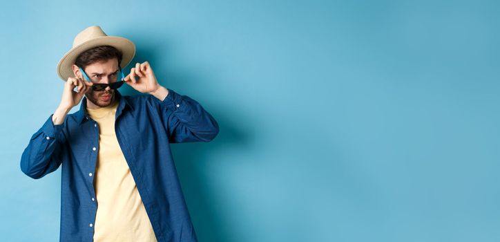 Portrait of guy on vacation staring aside with shocked and cringe face, take-off sunglasses and look at empty space on blue background.