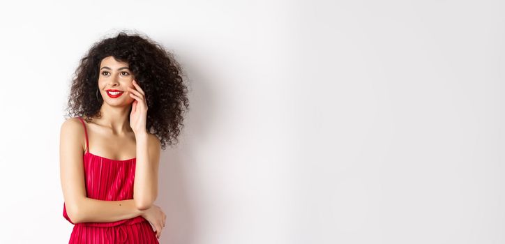 Elegant and romantic woman in red dress and makeup, smiling and touching her face, looking aside with happy expression, white background.