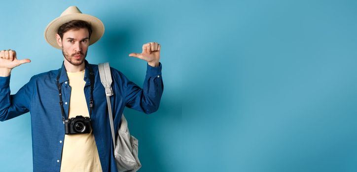 Confident handsome guy in summer hat, pointing at himself with bragging look, going to travel on holidays, holding backpack and camera, blue background.