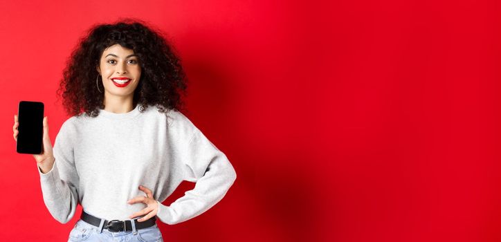 Attractive smiling woman showing empty smartphone screen and looking happy, advertising online store or application, standing against red background.