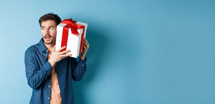 Valentines day. Curious boyfriend holding gift box near ear, trying to guess whats inside surprise present, standing over blue background.