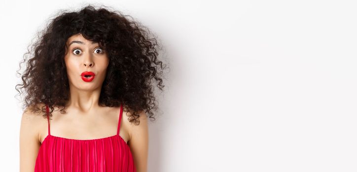 Close-up of surprised caucasian woman with red lips and curly hairstyle, saying wow and looking amazed, standing in red dress over white background.