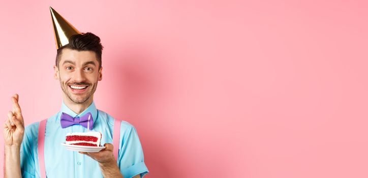 Holidays and celebration concept. Happy young man enjoying birthday party, wearing cone hat and cross fingers, making wish on bday cake with candle, pink background.