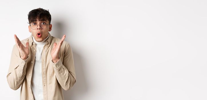 Portrait of surprised hipster guy in glasses checking out awesome promo, say wow and gasping amazed, raising hands up with disbelief, standing on white background.