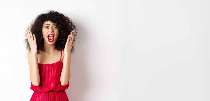 Woman scream in panic, wearing red dress and shouting at camera with anxious face, standing over white background. Copy space