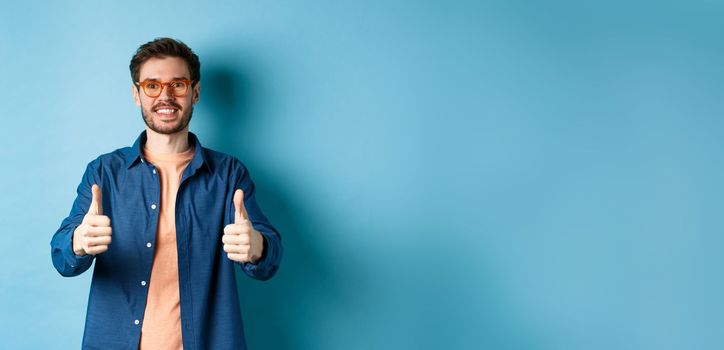 Happy young man in glasses showing thumbs up, recommending optician store, standing on blue background.