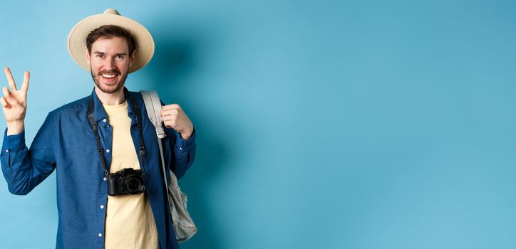 Happy handsome man taking photo on summer vacation, showing peace sign and smiling, wearing straw hat and holding tourist backpack, blue background.