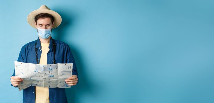 Covid-19, pandemic and travel concept. Young man in medical mask reading tourist map, searching for route to sightseeings, standing on blue background.