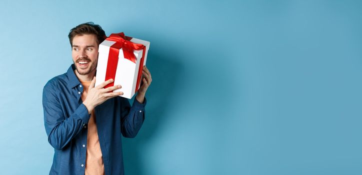 Valentines day. Smiling man guessing what inside gift box, got present from lover girlfriend, standing against blue background. Copy space