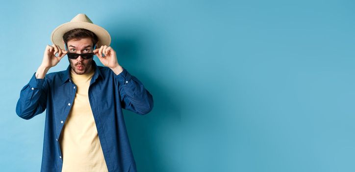 Portrait of tourist takes-off sunglasses and saying wow, staring impressed at camera, checking out cool thing, standing in summer hat on blue background.