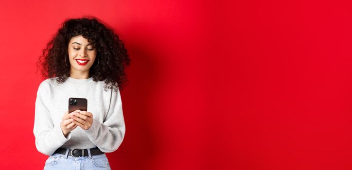 Smiling european woman reading mobile phone screen, texting a message on smartphone, standing in sweatshirt against red background.