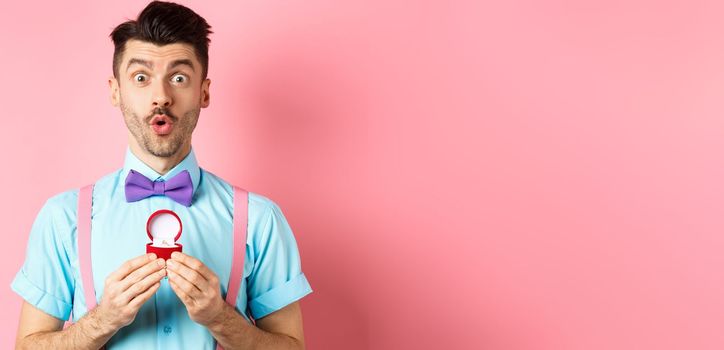 Valentines day. Funny man with moustache and bow-tie, looking excited and showing engagement ring, asking girlfriend to marry him, standing over pink background.